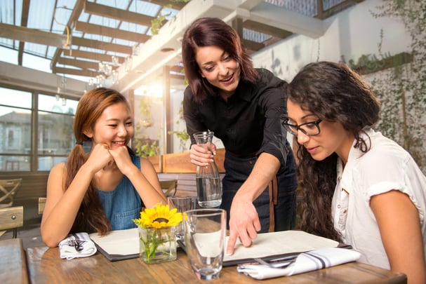 A server holding a carafe points to something on a menu while a second customer looks on. 