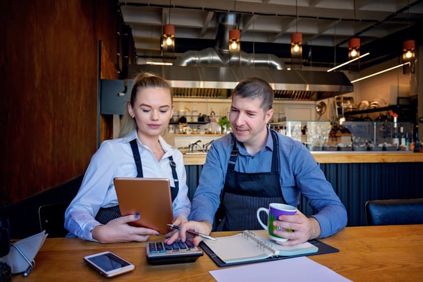 Two people wearing cooks' aprons sit at a restaurant table with paperwork and a calculator. 