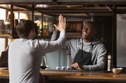 A smiling man in an apron standing behind a counter high-fives a customer. 
