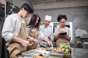A chef looks on as two people work at a kitchen counter and a third takes notes. 