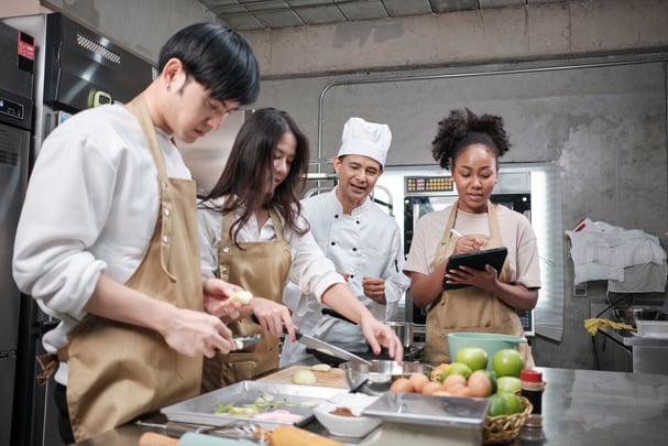 A chef looks on as two people work at a kitchen counter and a third takes notes.  