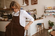 A long-haired man standing in a cafe puts on a brown apron. 