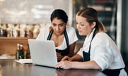 Two women wearing aprons sit in a restaurant working on a laptop computer. 