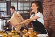 A smiling woman wearing a an apron holds a cardboard container over a display of rolls and pastries. 