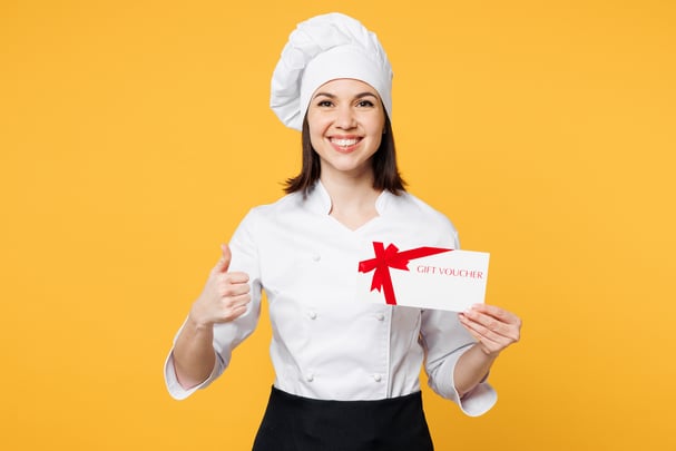 A woman dressed as a chef holds a gift card in one hand and gives a thumbs-up gesture with the other hand. 
