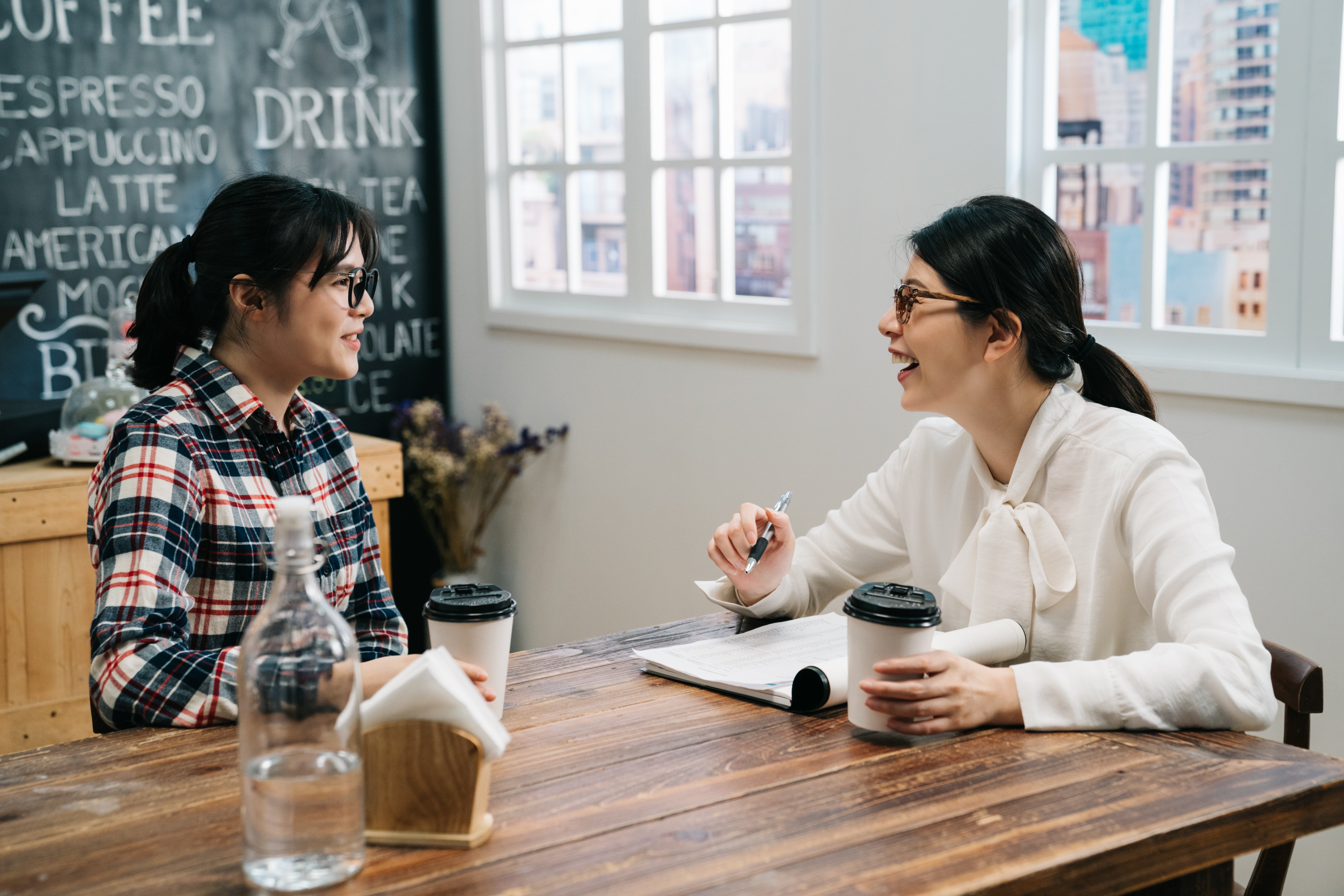 Two women talk at a table in a coffee shop; one has a pen in her hand and a notebook in front of her. 