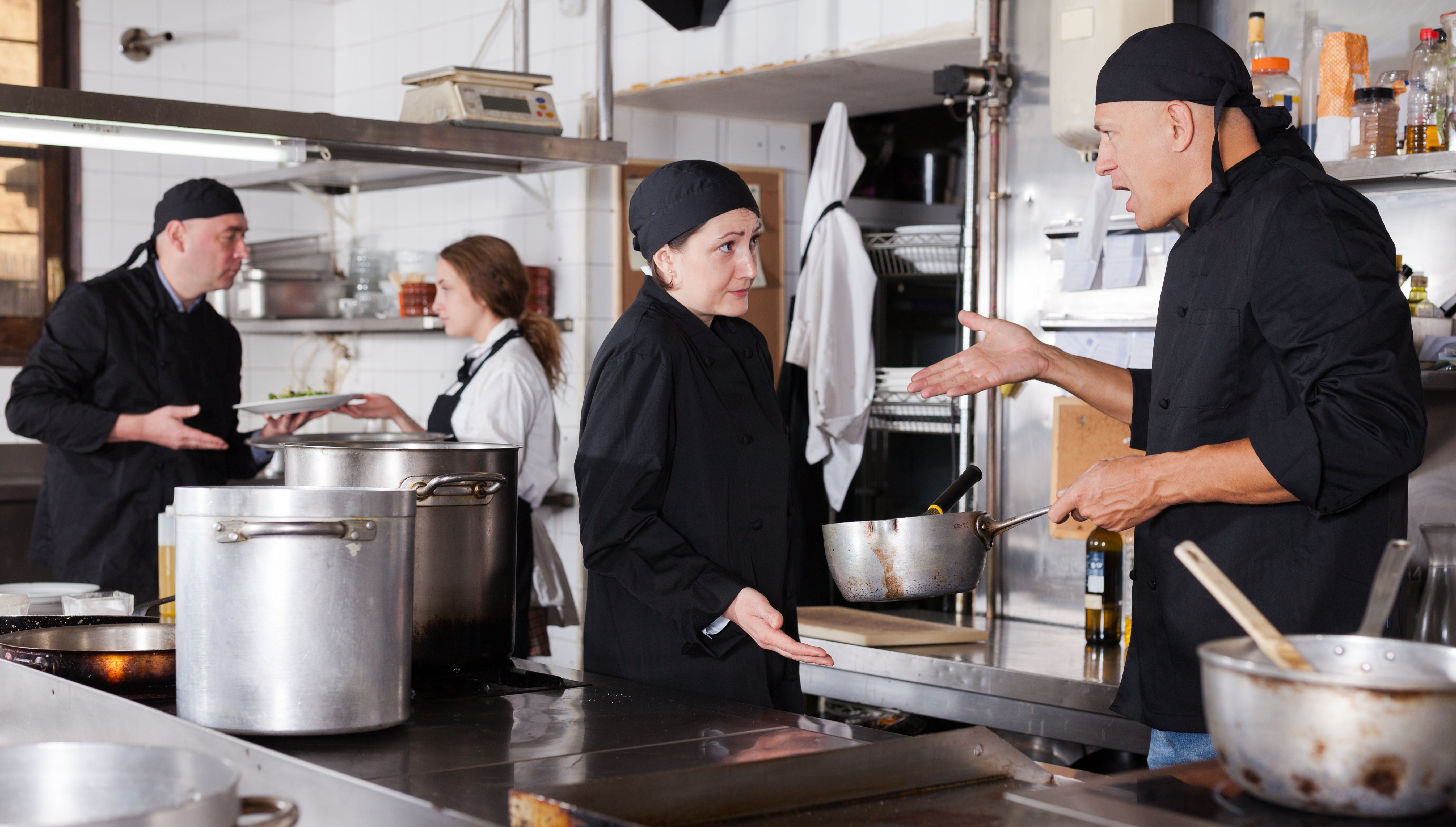 One cook yells at another, while in the background a third cook argues with a server. 