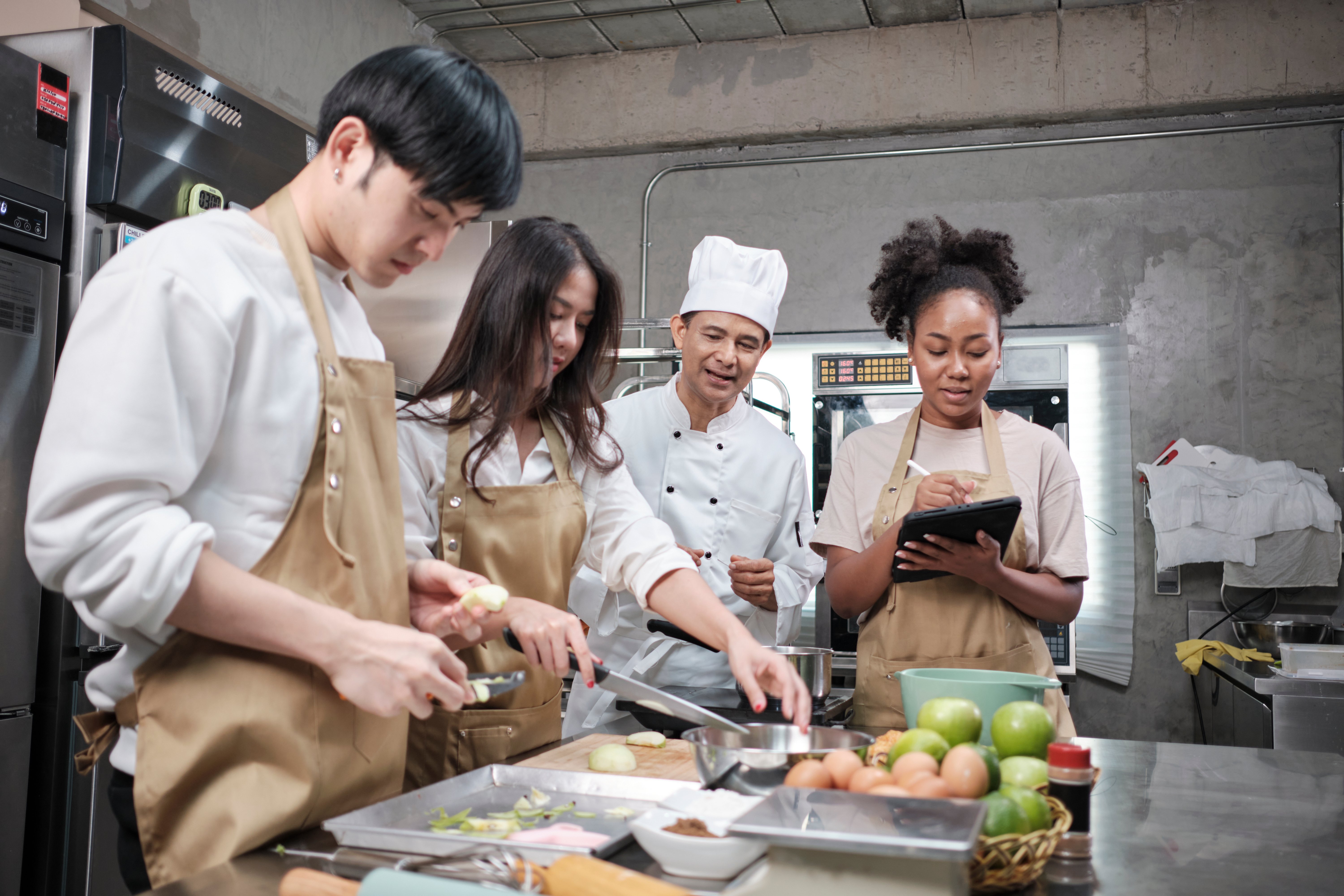 A chef looks on as two people work at a kitchen counter and a third takes notes. 