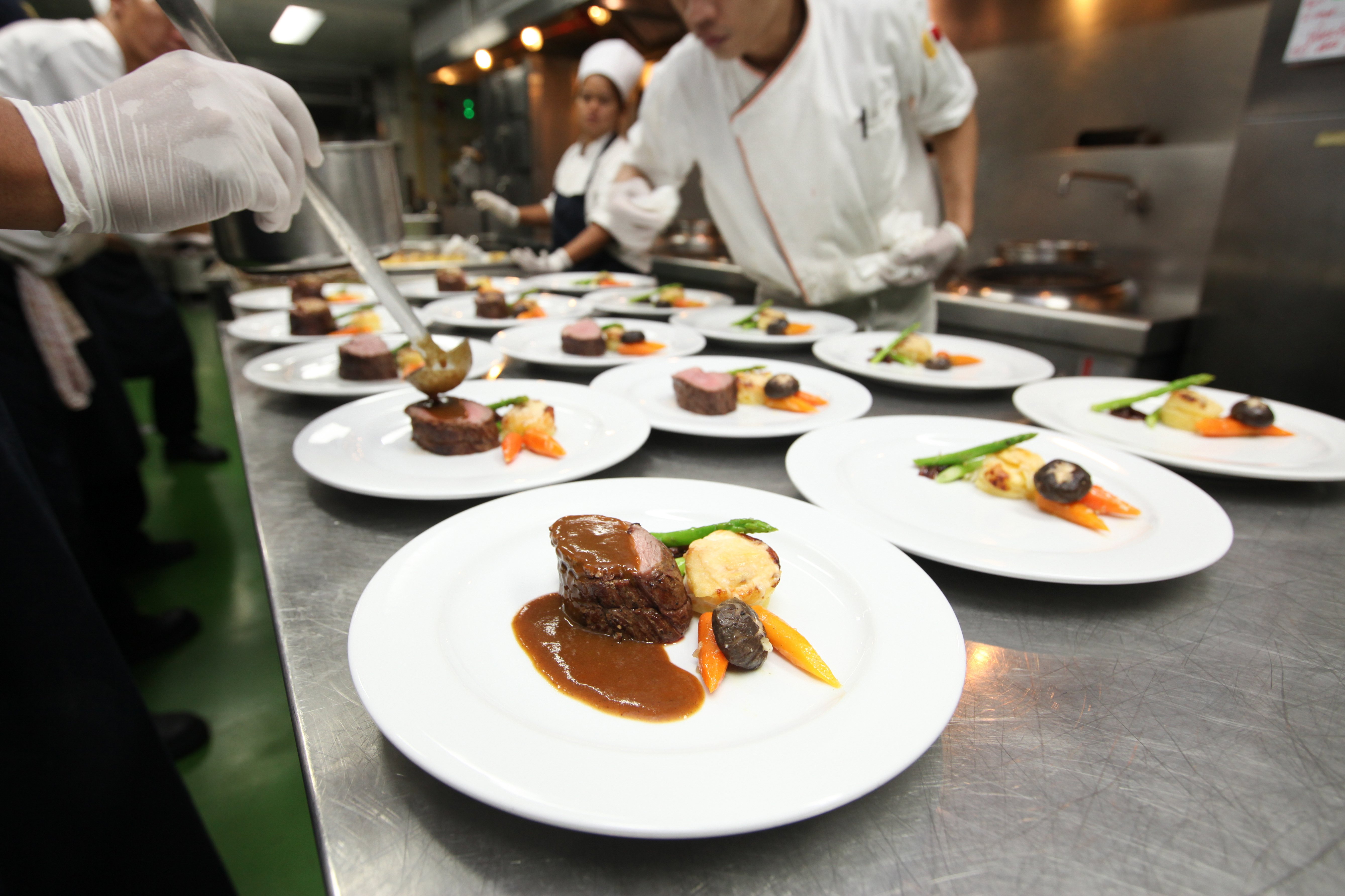 Workers add food to an array of plates on a restaurant kitchen counter.